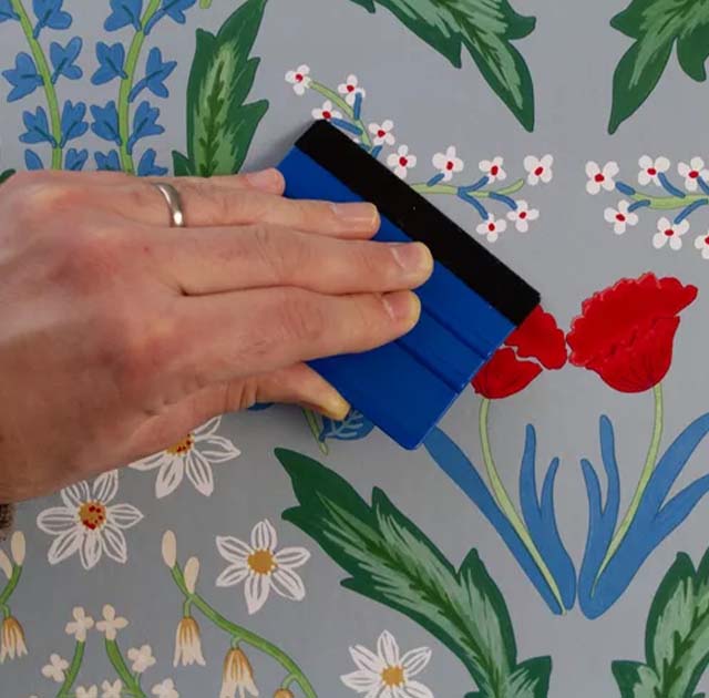 a-hand-smoothing-vibrant-floral-wallpaper-with-a-blue-tool-highlighting-intricate-blue-wallpaper-textures A hand smoothing vibrant floral wallpaper with a blue tool, highlighting intricate blue wallpaper textures.