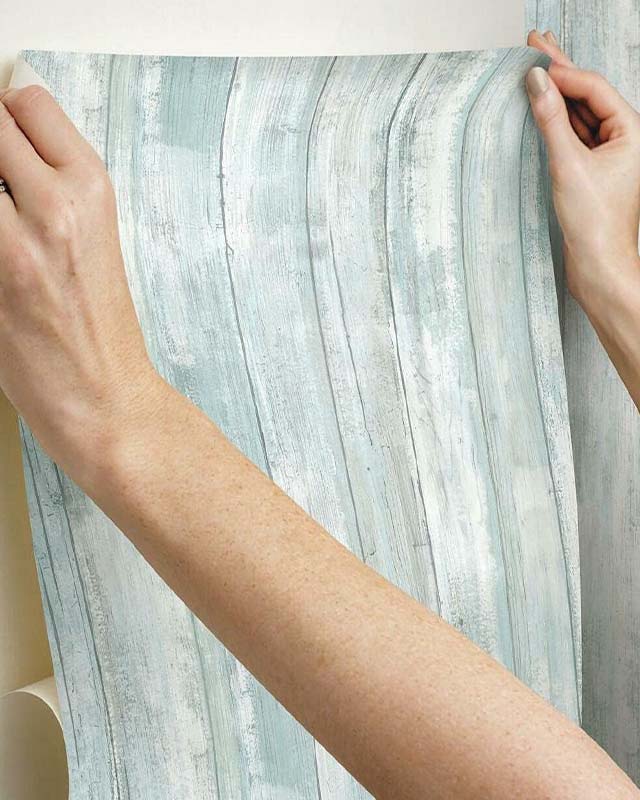 a-person-applies-a-light-blue-wood-textured-wallpaper-showcasing-current-blue-wallpaper-trends A person applies a light blue, wood-textured wallpaper, showcasing current blue wallpaper trends.