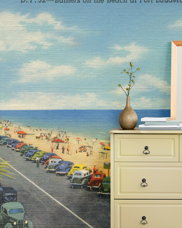 A room with vintage-style blue wallpaper trends featuring a beach scene, paired with a cream-colored dresser, vase, and books