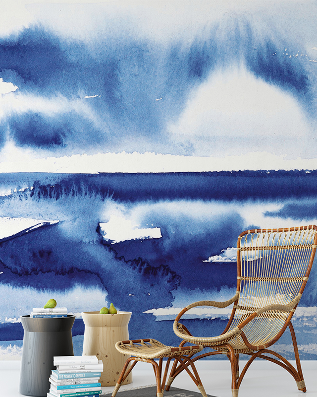 A watercolor-style mural depicting an abstract ocean scene in deep blue tones, showcasing blue wallpaper trends, with a wicker chair and side tables stacked with books in the foreground