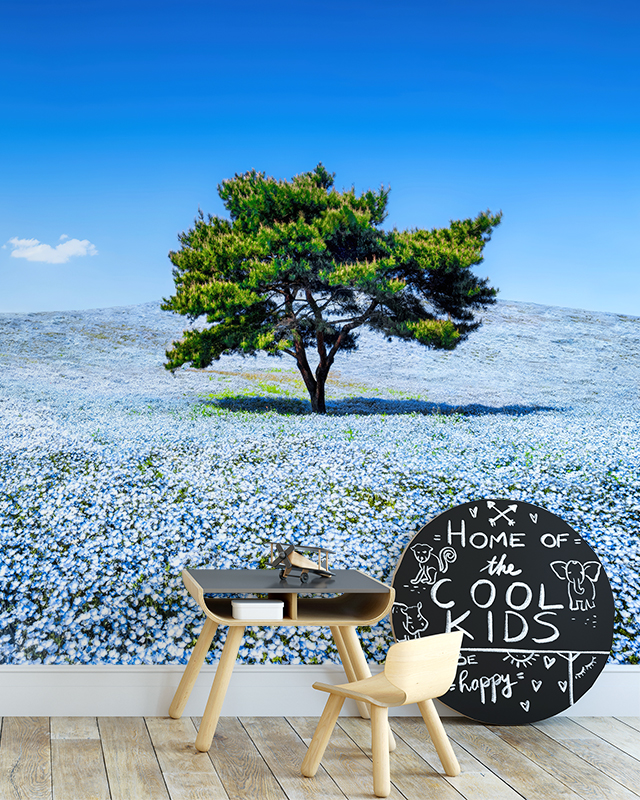 Kids' room with blue wallpaper trends featuring a scenic mural of a tree in a field of blue flowers under a bright blue sky, a small desk, and a chair