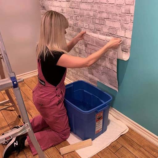 A-woman-in-pink-overalls-aligns-wallpaper-on-a-wall-during-Wallpaper-Installation-with-tools-and-water-nearby A woman in pink overalls aligns wallpaper on a wall during Wallpaper Installation, with tools and water nearby.