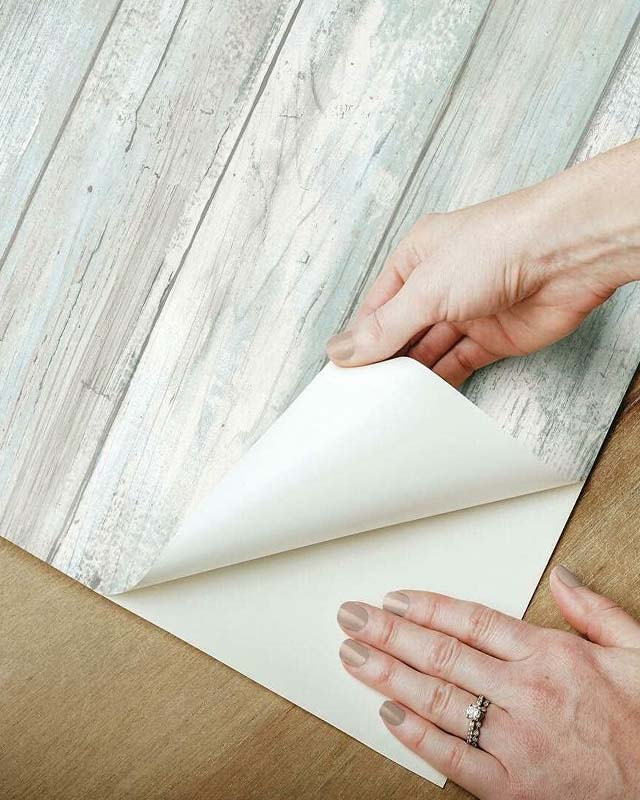 Hands-peeling-back-adhesive-backing-during-the-Wallpaper-Installation-process-on-a-wooden-surface. Hands peeling back adhesive backing during the Wallpaper Installation process on a wooden surface.