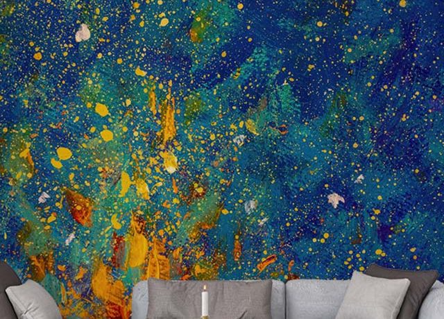Abstract wallpaper featuring a vibrant, splattered paint design in blue, yellow, and orange hues behind a modern sofa, ideal for Abstract Wallpaper for Accent Walls.
