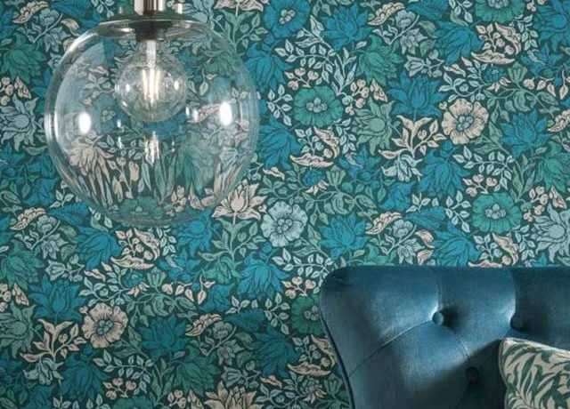 A glass pendant light hangs in front of floral William Morris wallpaper with modern art and a blue velvet chair.