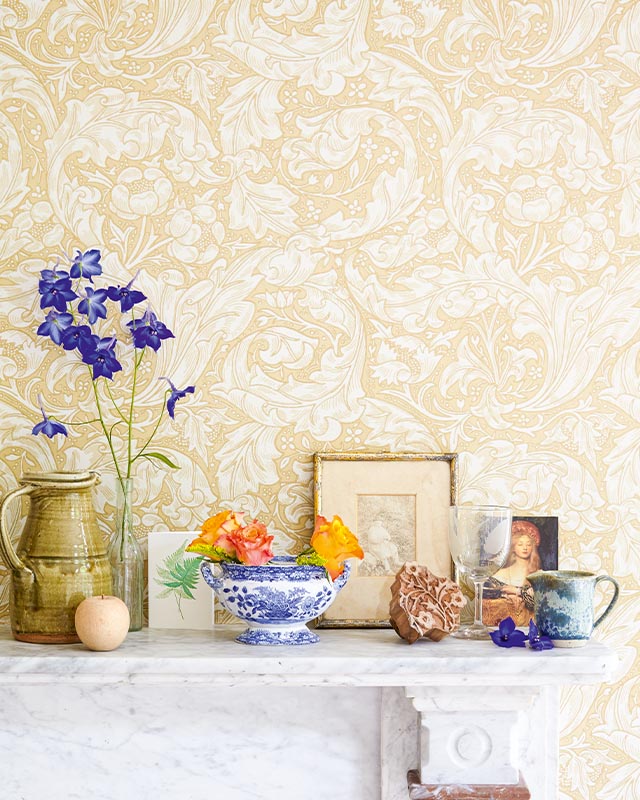 a-mantel-decorated-with-eclectic-items-against-beige-floral-william-morris-wallpaper-with-modern-art-inspired-accents A mantel decorated with eclectic items against beige floral William Morris wallpaper with modern art-inspired accents.