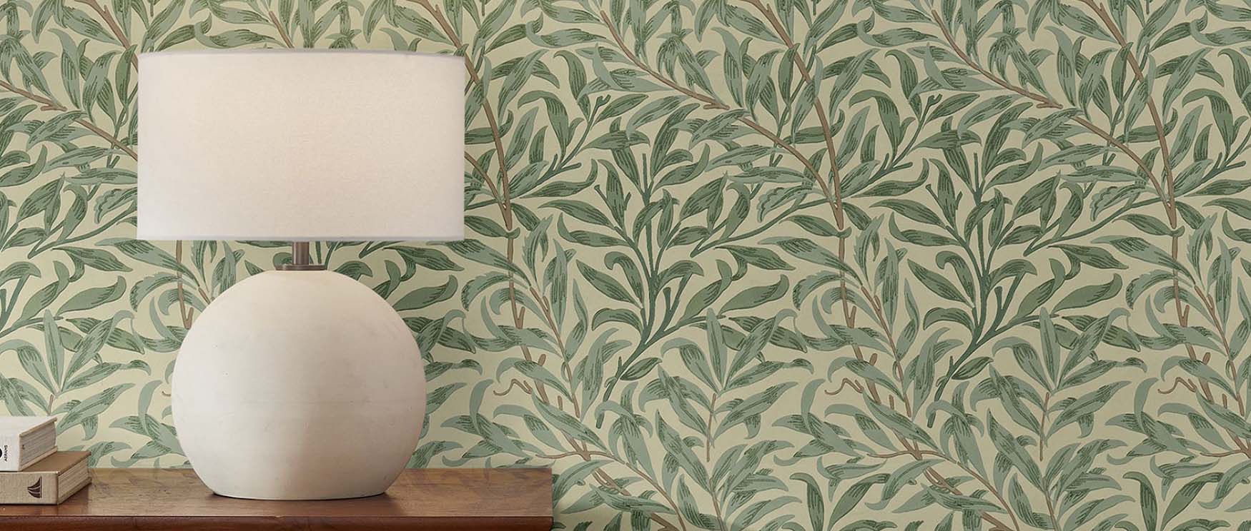 a-room-with-william-morris-wallpaper-patterns-featuring-green-leaves-and-a-large-round-table-lamp-on-a-wooden-surface A room with William Morris wallpaper patterns featuring green leaves and a large round table lamp on a wooden surface.