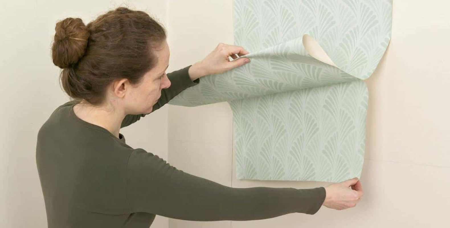 a-woman-installs-peel-and-stick-wallpaper-with-a-green-leaf-pattern-smoothing-it-onto-the-wall A woman installs peel and stick wallpaper with a green leaf pattern, smoothing it onto the wall.