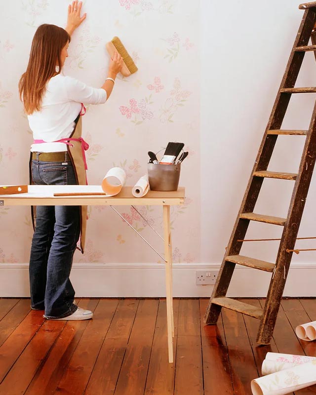 a-woman-smooths-floral-peel-and-stick-wallpaper-on-a-wall-with-a-brush-tools-nearby-for-installing-peel-and-stick-wallpaper A woman smooths floral peel and stick wallpaper on a wall with a brush, tools nearby for installing peel and stick wallpaper.