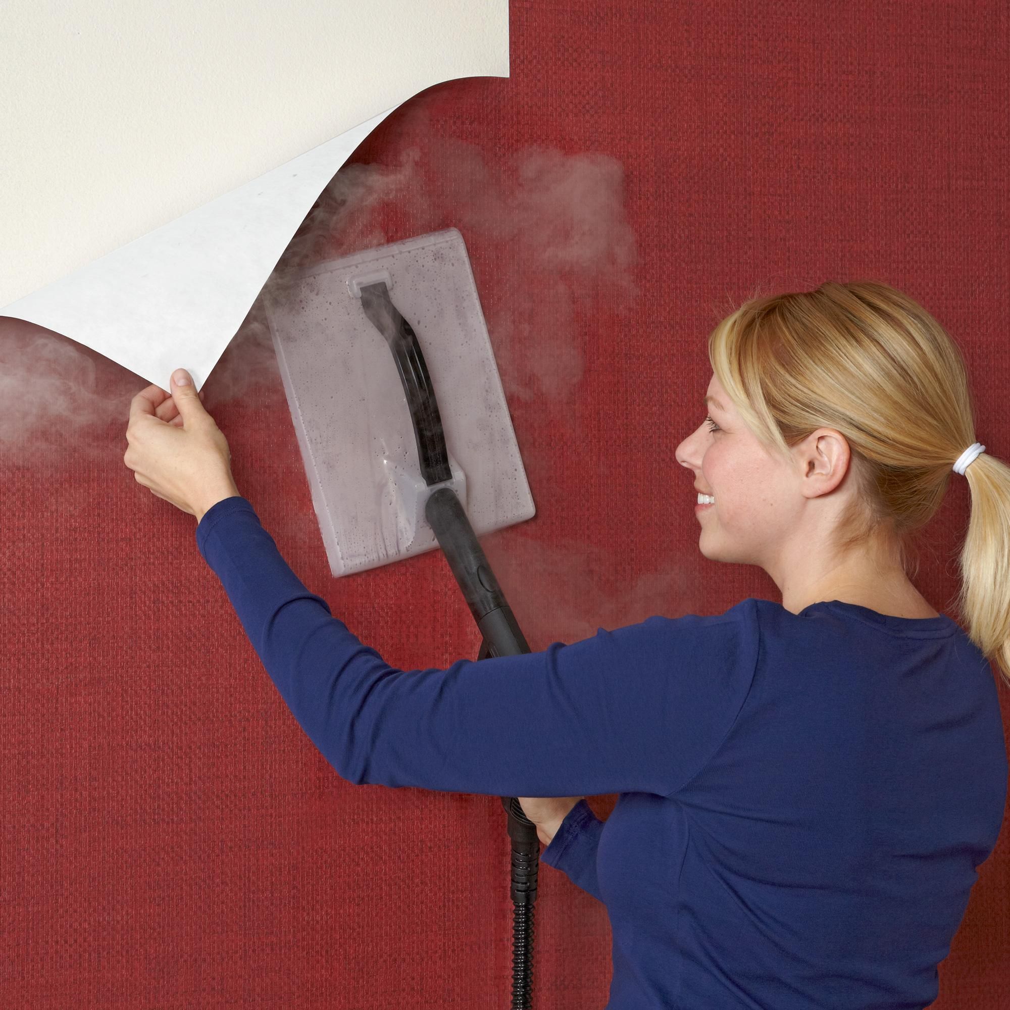a-woman-uses-a-steamer-to-remove-peel-and-stick-wallpaper-from-a-red-wall-during-installation-preparation A woman uses a steamer to remove peel and stick wallpaper from a red wall during installation preparation.