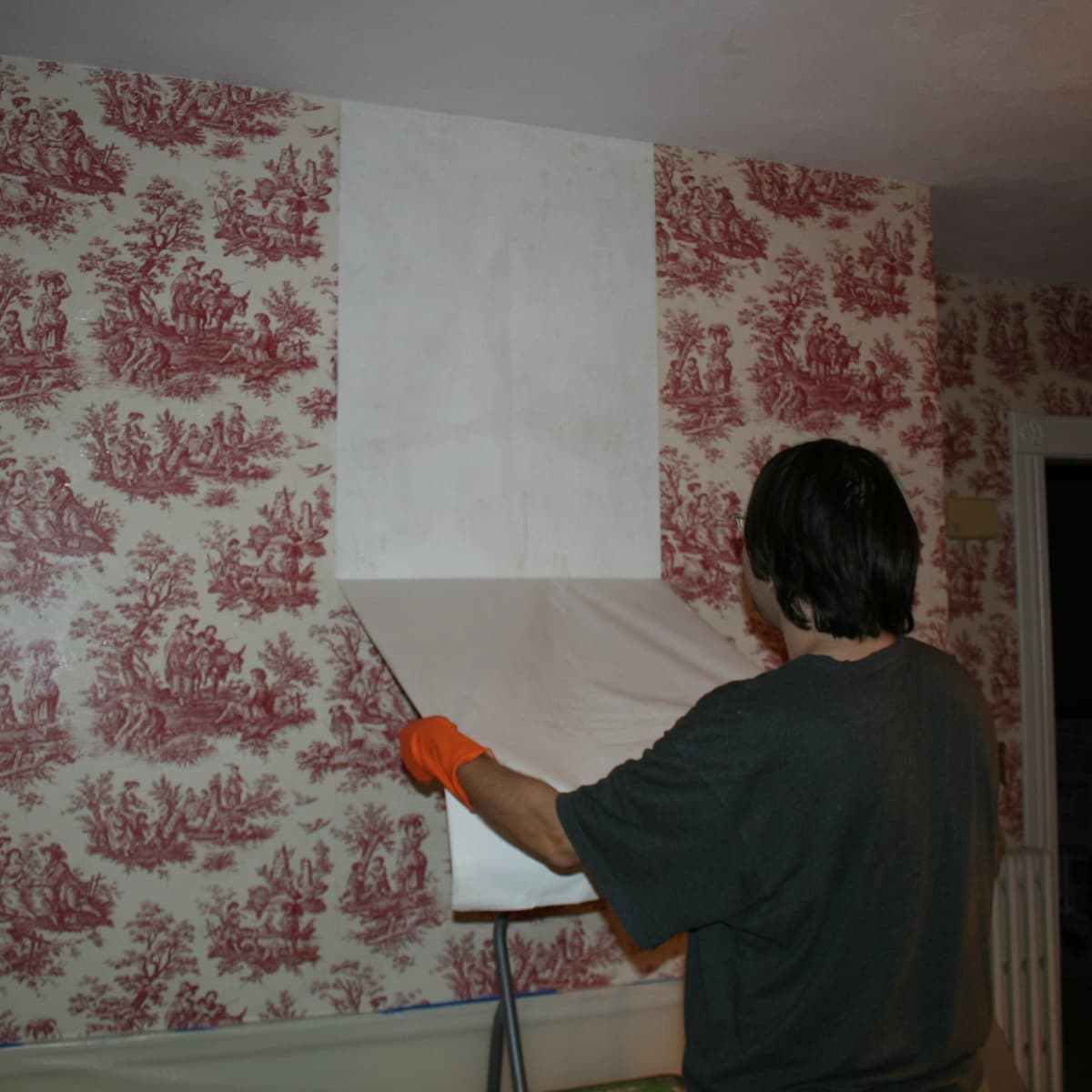 person-removing-red-patterned-peel-and-stick-wallpaper-for-rental-space-revealing-a-blank-white-wall-underneath Person removing red-patterned peel and stick wallpaper for rental space, revealing a blank white wall underneath.
