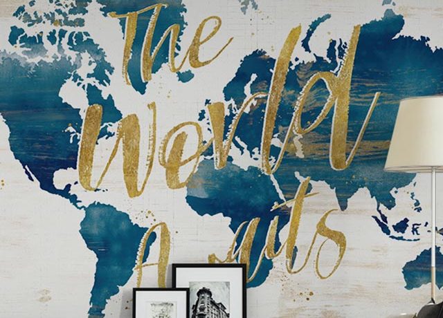 World map mural with "The World Awaits" text, alongside framed pictures and a lamp, featuring peel and stick wallpaper for rental space.