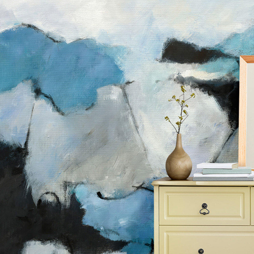Abstract painting with blue wallpaper behind a yellow dresser featuring a vase and books in a modern room. 