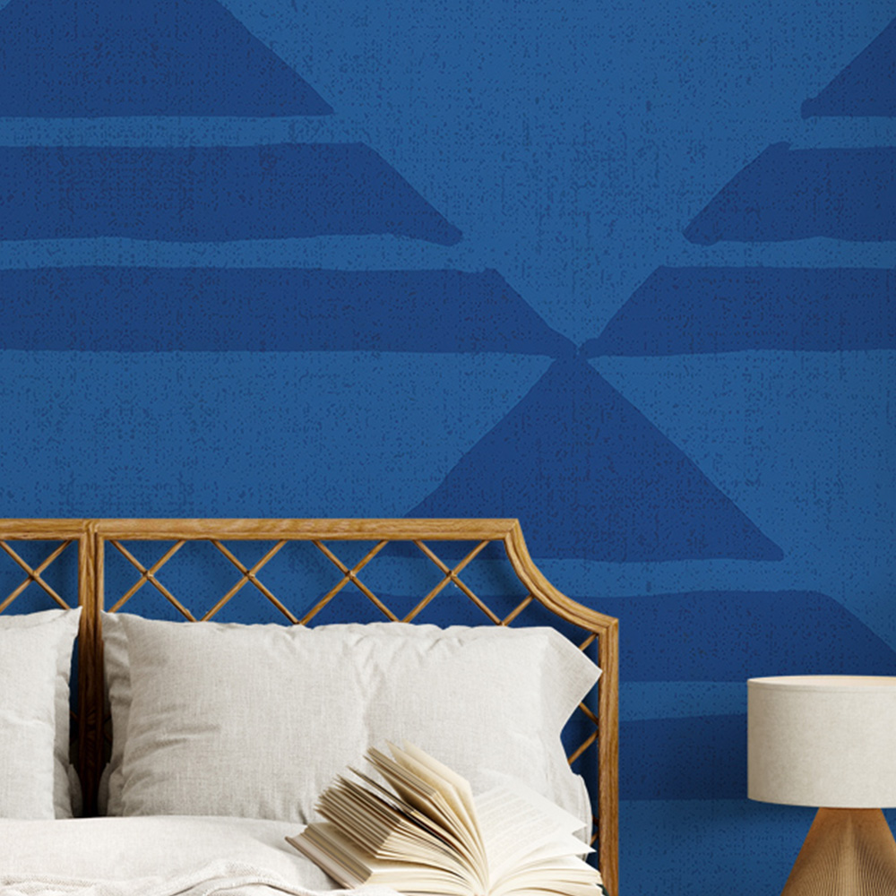 Geometric pattern in blue wallpaper behind a bed with a beige headboard and a side lamp in a cozy bedroom. 