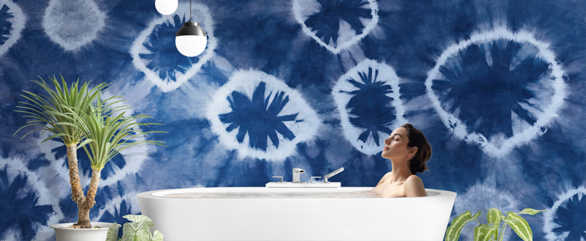 Relaxing bathtub scene with tie-dye patterned blue wallpaper, tropical plants, and modern pendant lighting. 